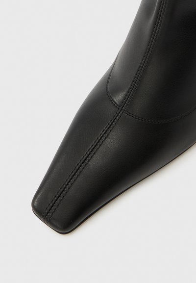Melns ādas potīšu zābaks ar smailu purngalu un redzamām šuvju detaļām. Gluda tekstūra ar elegantu, mūsdienīgu dizainu.