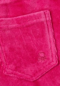 Zbliżenie na jasnoróżową tkaninę sztruksową, której elementem jest przeszywana kieszeń z małym haftowanym symbolem w prawym dolnym rogu.