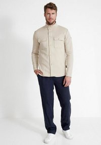 Beige Langarm-Hemdjacke mit zwei Fronttaschen, Knopfverschluss und Stehkragen, kombiniert mit marineblauen Hosen und weißen Sneakers.