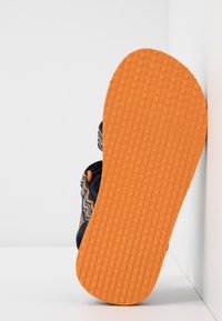 Sanda je vybavena texturovanou oranžovou gumovou podrážkou a námořnickým svrškem s modrými a oranžovými cik-cak vzory; viditelné detaily nastavitelného pásku.