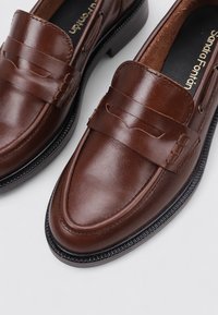 Mocasines de cuero marrón con un acabado suave, que cuentan con un diseño de correa tradicional, punta redondeada y una suela de goma negra texturizada.