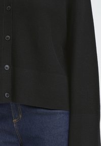 Fekete kardigán texturált kötött mintával, gombos elülső résszel és bordás szegéllyel. Sötétkék farmerrel viselve, varrási részletekkel.