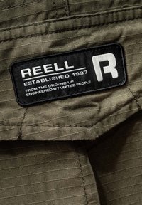 Olive green fabric with a textured pattern; features a black label with white text stating "REELL ESTABLISHED 1997" and a logo.