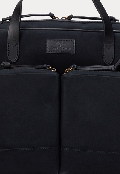 Saco de lona preto com detalhes em pele, apresentando dois bolsos frontais e um rótulo de marca. Textura suave e detalhes em metal dourado.