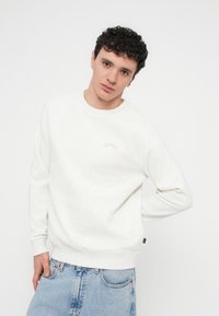 Sweatshirt branca de gola redonda com punhos e bainha canelados, apresentando um detalhe sutil do logotipo no peito. Usada com jeans de ganga azul.