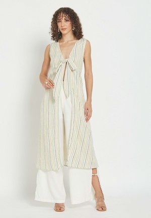 Ženska z kodrastimi lasmi, ki nosi brezrokaven dolg teliran telovnik s črtami, zavezano spredaj, bele hlače širokih hlačnic in prozorne sandale z odprtimi prsti, stoji ob beli steni.