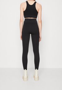 Femme portant un soutien-gorge de sport noir Puma et un legging taille haute, se tenant dos au spectateur, avec des bottes blanches chunky devant un fond uni.