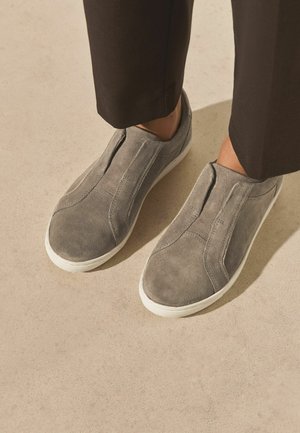 Pilkos spalvos zomšos slip-on batai su baltais pado padėtais prie juodų trumpintų kelnių ant šviesiai kreminės grindų.