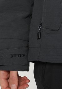 Gros plan d'une manche de veste sombre avec le logo "BURTON" brodé près du poignet et une main partiellement visible en dessous.