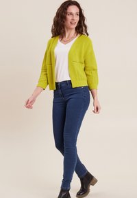 Cardigan lavorato a maglia di un vivace giallo con maniche a tre quarti e design aperto, indossato sopra una maglietta bianca e jeans blu, abbinato a stivali neri alla caviglia.