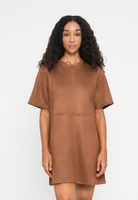 Vrouw met lang krullend haar, gekleed in een korte mouw, boven de knie, bruine jurk, staande voor een effen witte achtergrond.