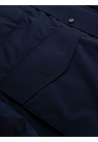 Detail kapsy námořnickomodré bundy s klopou a viditelným stehem, která má nad kapsou jeden černý knoflík.
