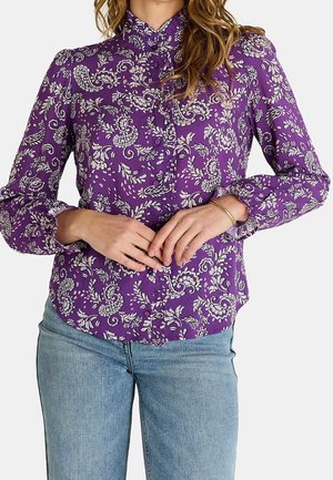 Kvinde iført en lilla bluse med lange ærmer med hvide blomstrede paisley mønstre og lyseblå jeans, hænder samlet ved taljen.
