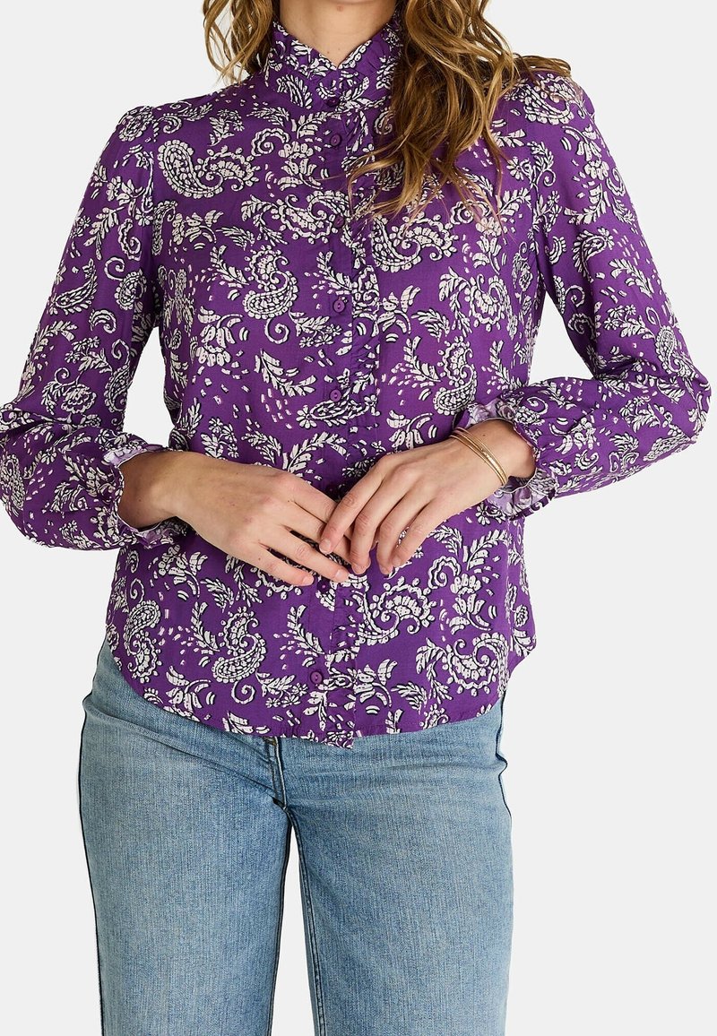 Femme portant un chemisier violet à manches longues avec des motifs floraux blancs paisley et un jean bleu clair, les mains jointes à la taille.