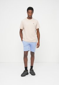 Uomo in piedi con le mani nelle tasche, indossa una camicia beige a maniche corte con motivo, pantaloncini azzurri, calzini neri e scarpe da ginnastica nere su sfondo bianco.