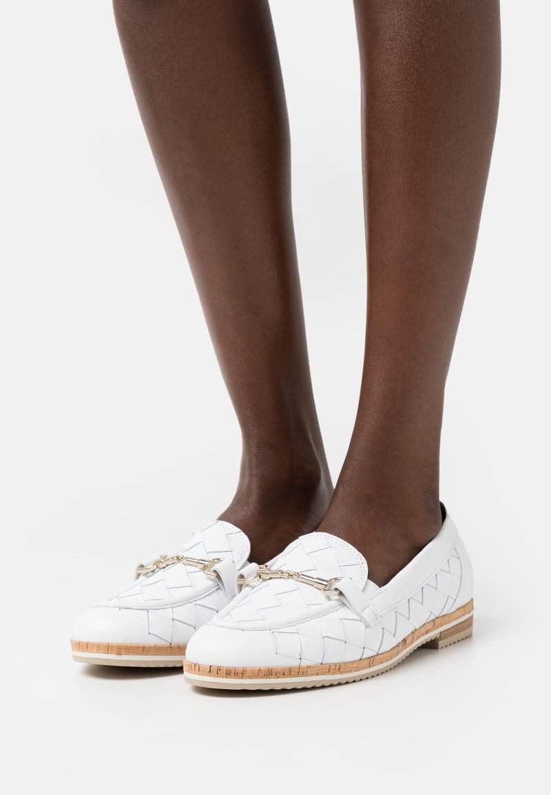 Pieds portant des mocassins en cuir tressé blanc avec détail en chaîne dorée et garniture en liège, posés sur une surface blanche unie.