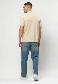 Egy férfi hátulról, bézs rövid ujjú pólót, kék farmert és fehér sportcipőt visel egy egyszínű világos háttér előtt.