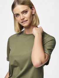 Žena s plavom bob frizurom koja nosi maslinasto zelenu majicu, gleda u kameru dok joj je desna ruka položena na lijevo rame.