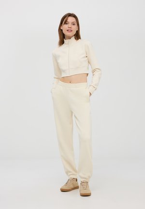 Felpa corta zip-up color crema con tasca frontale, abbinata a pantaloni sportivi tapered coordinati e sneaker color marrone chiaro. Texture liscia.