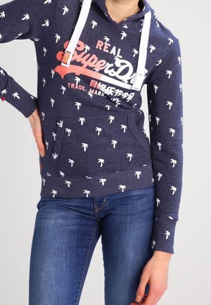 Marineblauwe hoodie met wit palmbomenpatroon, witte trekkoorden, "Real Superdry" logo in koraal en wit, gedragen met blauwe spijkerbroek, hand op heup.