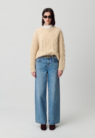 Maglione a trecce crema, jeans larghi azzurri, stivali alla caviglia marroni e occhiali da sole scuri; presenta motivi testurizzati e vestibilità rilassata.