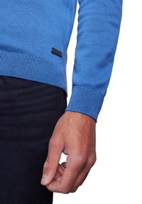 Blauer Pullover mit geripptem Saum und Bündchen, aus einem glatten Stoff gefertigt. Mit einem kleinen schwarzen Logolabel an der Seite.