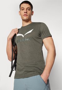 Olijfgroen t-shirt met korte mouwen, gemaakt van zachte stof, met een wit vogellogo en de tekst "SALEWA" gedrukt op de voorkant.