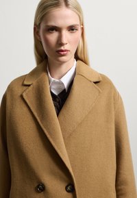 Cappotto di lana color cammello con design doppiopetto, colletto a rever e due bottoni neri; indossato sopra una camicia bianca.