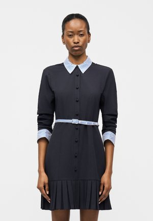 Γυναίκα που φοράει μαύρο φόρεμα με μακριά μανίκια και γιακά, μανσέτες και ζώνη με μπλε και λευκές ρίγες, στέκεται μπροστά σε απλό φόντο.