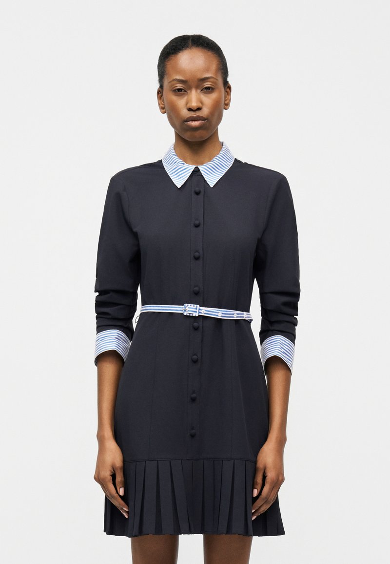 Ženska v črni obleki z dolgimi rokavi, z modro-belim črtastim ovratnikom, manšetami in pasom, stoji pred enobarvnim ozadjem.