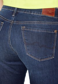 Dark blue denim jeans with a smooth texture, featuring orange stitching, two back pockets, and a brown leather brand patch at the waistband.