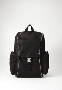 Mochila de tela negra con cierre de cremallera, dos bolsillos laterales, herrajes plateados y asa superior. En el frente presenta un logo y detalle de hebilla.