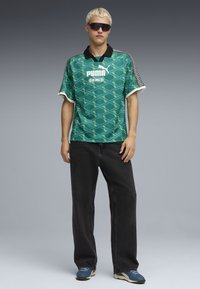 Polo de manga curta com padrão verde e colarinho preto, combinado com jeans pretos largos. A camisa apresenta uma marcação ousada e detalhes contrastantes.