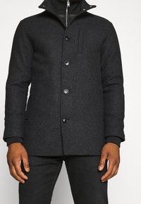 Uomo che indossa un cappotto di lana grigio scuro con bottoni sopra una giacca nera con zip, abbinato a pantaloni neri, su uno sfondo chiaro e semplice.