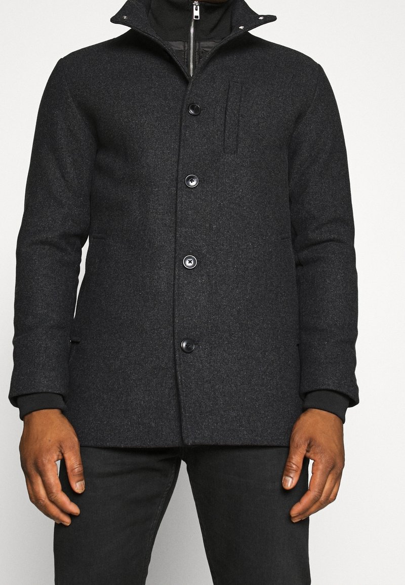 Uomo che indossa un cappotto di lana grigio scuro con bottoni sopra una giacca nera con zip, abbinato a pantaloni neri, su uno sfondo chiaro e semplice.