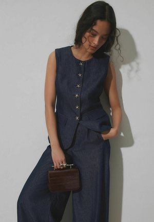 Jeansweste mit sternförmigen Knöpfen, figurbetontem Schnitt und ärmellosem Stil; kombiniert mit weiten Jeanshosen und einer kleinen dunkelbraunen Handtasche.