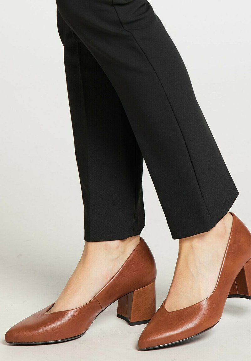 Décolleté in pelle marrone con punta affilata e design con ritagli, posizionati su un tacco a blocco spesso. Abbinati a pantaloni neri sartoriali.