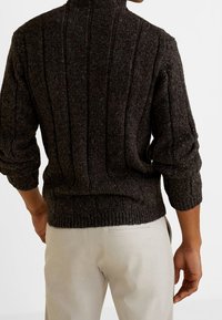 Pull en maille gris foncé avec motif côtelé vertical, col roulé et manches larges. Le matériau semble texturé et confortable. Porté avec un pantalon clair.