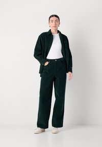 Dunkelgrünes Cordhemd und -hosen-Set, das einen lässigen Schnitt, einen Knopfverschluss und ein weißes T-Shirt darunter aufweist. Glatte Textur, lässiger Stil.