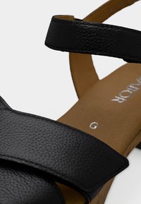 Μαύρο δερμάτινο σανδάλι με δύο φαρδιές λουράκια, υφασμένο επάνω μέρος, ταμπά πέλμα και φελλό σόλα. Περιλαμβάνει εμφανείς ραφές και διακριτική λεπτομέρεια λογότυπου.