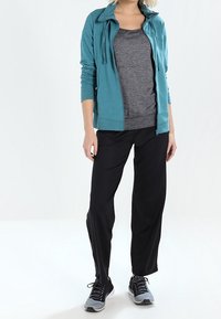 Persona vestida con una sudadera con cremallera color turquesa, camiseta gris oscuro, pantalones negros y zapatillas negras y grises, de pie contra un fondo blanco.