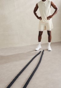Top beige senza maniche e pantaloncini indossati con sneakers bianche e calze. Due corde nere attorcigliate sono adagiati su un pavimento grigio chiaro.