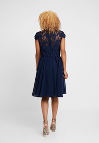 Robe bleu marine avec un corsage en dentelle ajusté et de courtes manches gigot, s'évasant en une jupe plissée. Le dos présente des détails complexes en dentelle florale.
