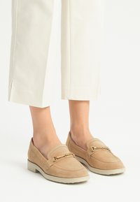 Beige mokasiner av semsket skinn med flat såle, prydet med en dekorativ metallkjede over framsiden og en glatt overflate.