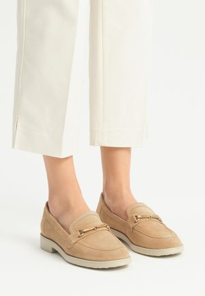 Mocassini beige in suede con suola piatta, caratterizzati da una catena metallica decorativa sulla parte anteriore e una finitura liscia.