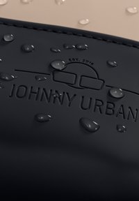 Fekete vízálló anyag, kiemelkedő "JOHNNY URBAN" logóval és "EST. 2016." felirattal. A felszínen lévő vízcseppek kiemelik a textúrát és a felületet.