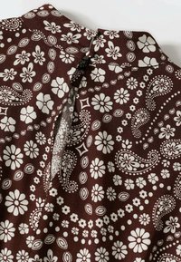 Braun gemusteter Stoff mit weißen floralem und Paisley-Designs. Der Kragen hat eine kleine Öffnung, die filigrane Details zeigt.