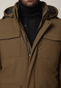 Brązowa ocieplana kurtka z zamkiem błyskawicznym, odpinanym kapturem, dwoma kieszeniami na piersi i gładką fakturą, noszona na czarnym golfie.