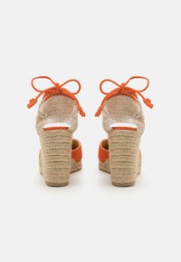 Zeppe espadrille arancioni con texture in juta intrecciata, schiena aperta e dettagli a fiocco. Tacchi spessi e texturizzati per stabilità e comfort.