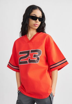 Vrouw met zwarte zonnebril en een los rood shirt met nummer 23 en mouwen met zwarte strepen, staand met handen in de zakken.